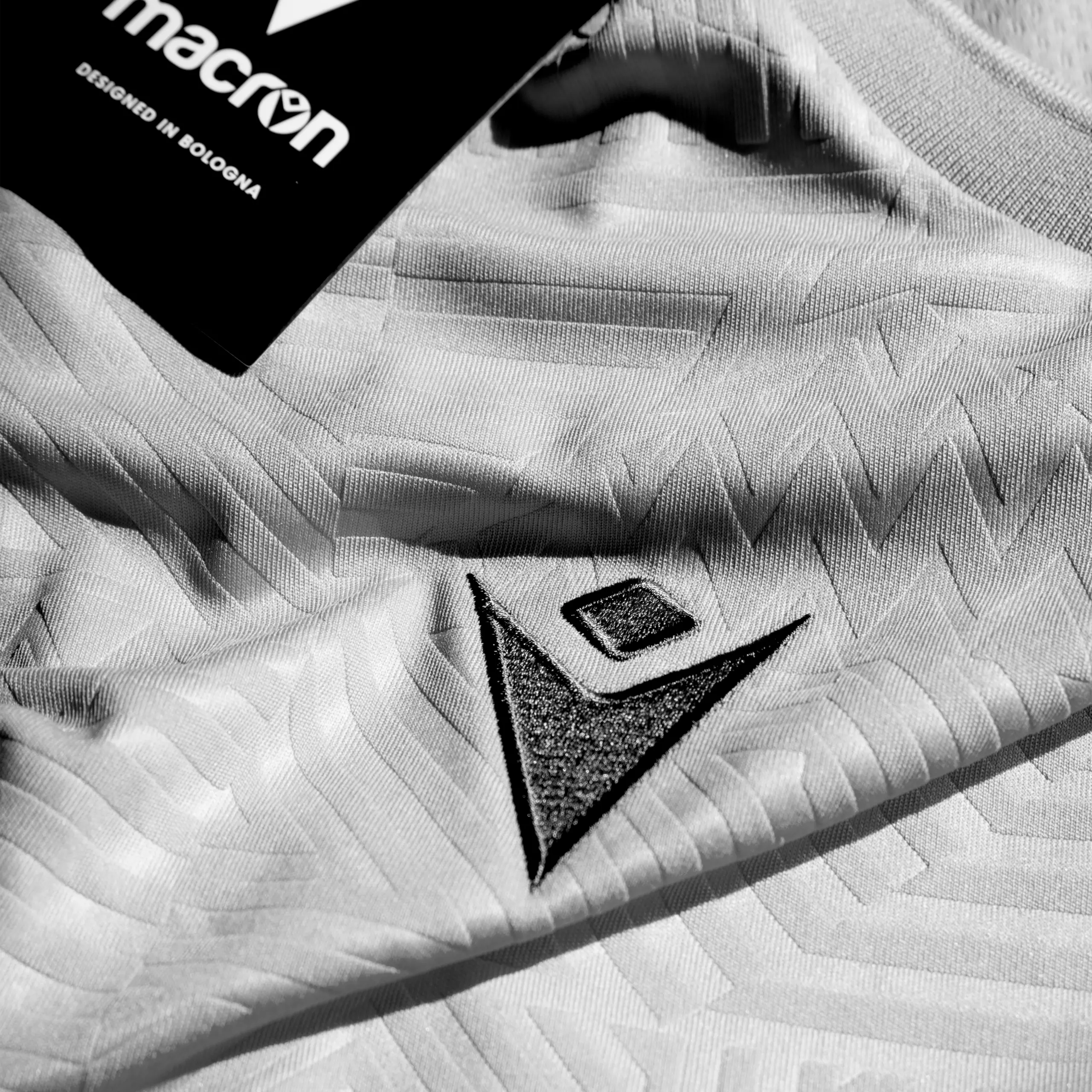 TShirt_Blue_Close_B&W_SQ Black and white close up of a football top with a stitch macron logo and macron card tag in background.